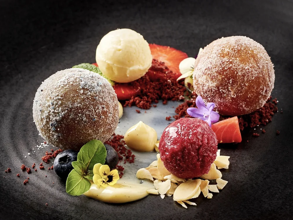 Hotel Salnerhof | 4 Sterne Superior in Ischgl Dessert mit zwei Puderzuckerbällchen, zwei Eiskugeln und essbaren Blumen auf schwarzem Teller