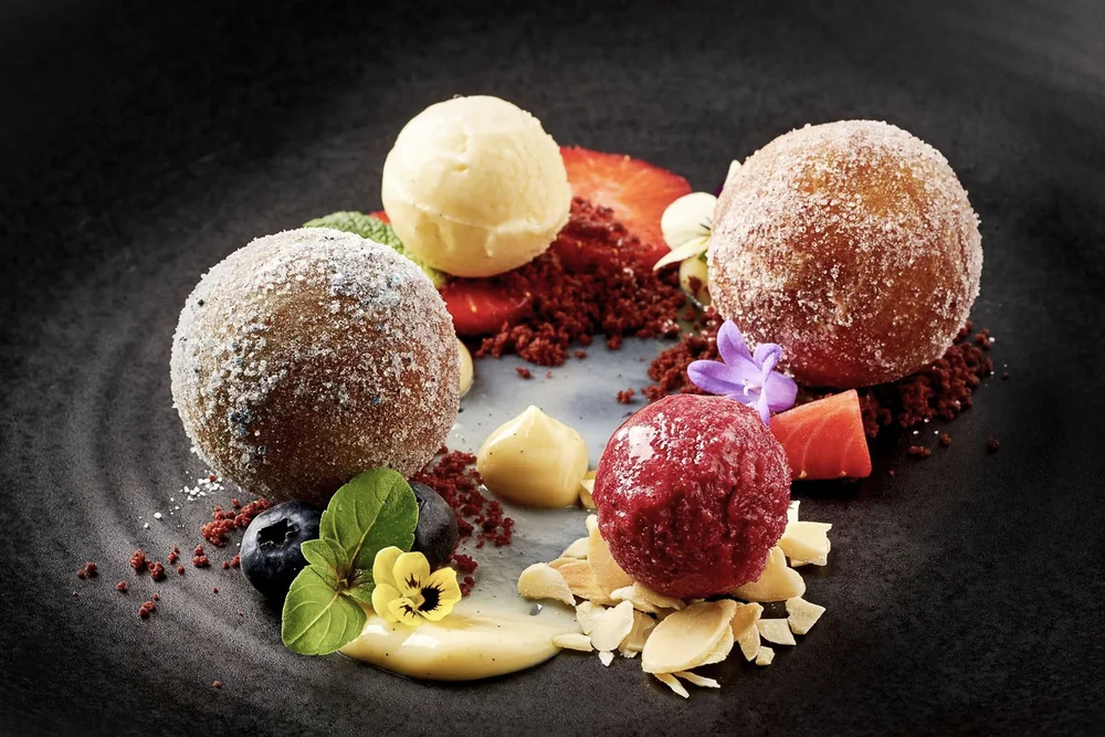 Dessert mit zwei Puderzuckerbällchen, zwei Eiskugeln und essbaren Blumen auf schwarzem Teller