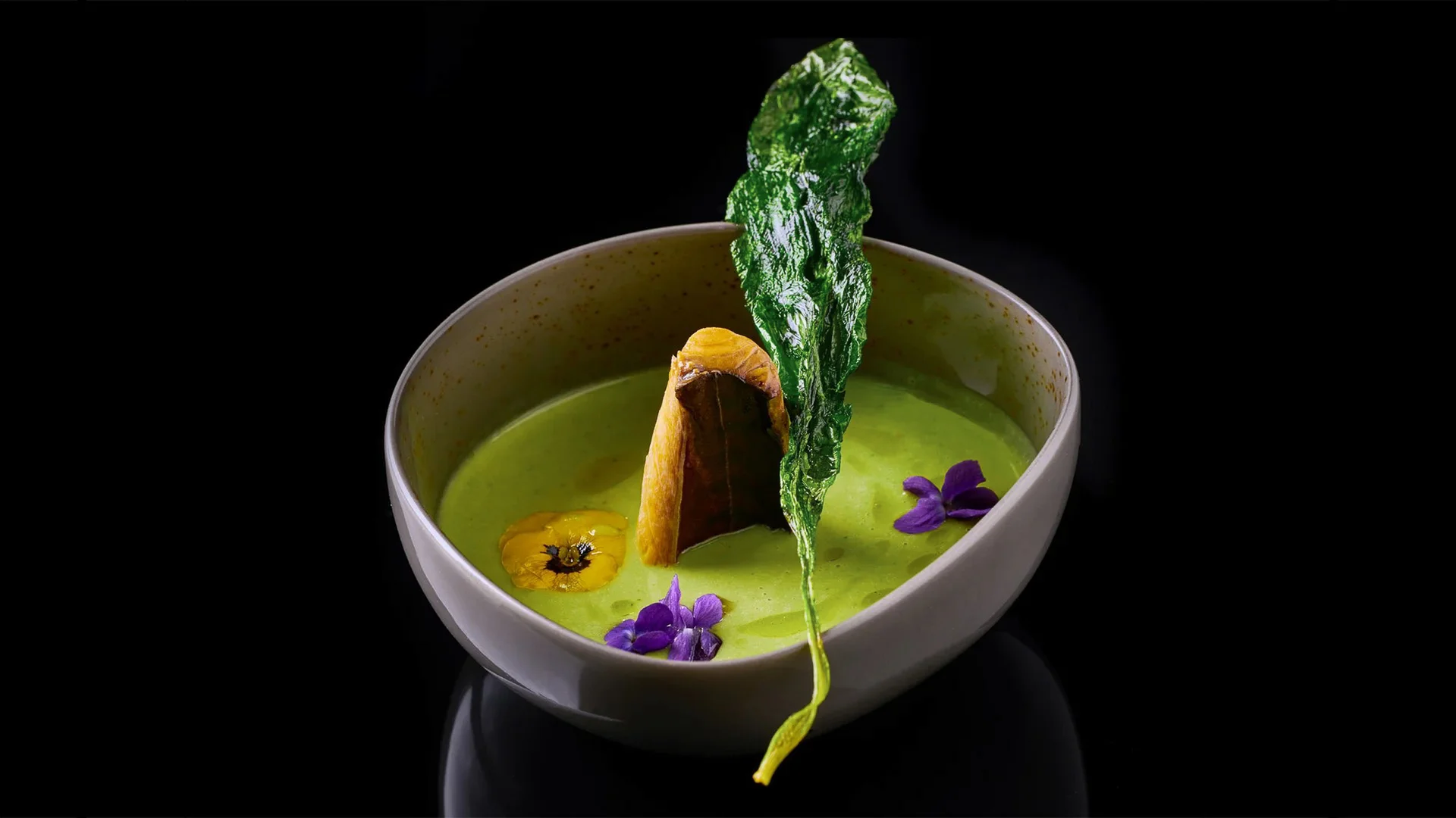 Grüne Suppe mit essbaren Blüten und knusprigem Blatt in weißer Schale